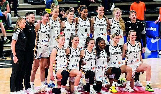 Auf dem Bild ist eine Mannschaft in weißen Trikots auf einem Basketballfeld zu sehen, die für ein gemeinsames Gruppenfoto posiert. Die Spielerinnen und Spieler sind in zwei Reihen angeordnet und werden von Mitgliedern des Trainer- oder Betreuerteams flankiert. Die Aufnahme entstand in einer Sporthalle und vermittelt den Eindruck einer offiziellen Teampräsentation nach einem Wettkampf oder Turnier.