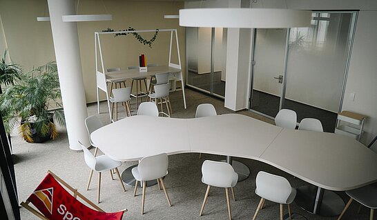 Ein moderner, heller Besprechungs- oder Coworking-Bereich mit einem großen geschwungenen weißen Konferenztisch und mehreren weißen Stühlen steht im Mittelpunkt des Raums. Im Hintergrund befinden sich weitere Arbeitsplätze mit einem Hochtisch, Glaswänden, Pflanzen und einer offenen, zeitgemäßen Bürogestaltung.