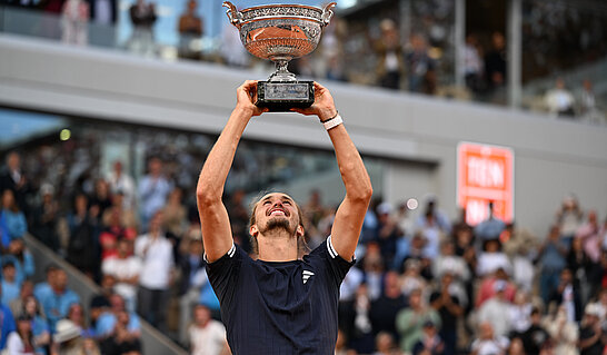 Eine Person auf einem Tennisplatz hält mit beiden Armen eine große silberne Trophäe über den Kopf. Im Hintergrund sind unscharfe Zuschauertribünen zu sehen, wodurch der Fokus klar auf der Person und dem Pokal liegt.