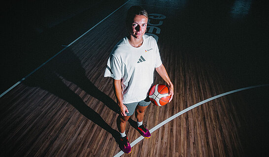 Die Aufnahme zeigt eine Person mit einem Basketball auf einem Hallenspielfeld aus erhöhter Perspektive. Die gezielte Beleuchtung, die markanten Spielfeldlinien und der lange Schatten verleihen dem Bild eine dynamische und fokussierte Wirkung.