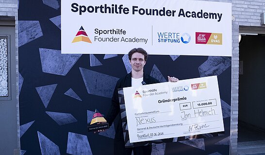 Eine Person steht vor einer großen Sponsorentafel der „Sporthilfe Founder Academy“ und hält einen übergroßen symbolischen Scheck sowie eine Trophäe in den Händen. Auf dem Scheck ist eine Gründerprämie über 15.000 Euro zu sehen, während im Hintergrund die Logos der Sporthilfe Founder Academy, der Werte Stiftung sowie weiterer Partner abgebildet sind.