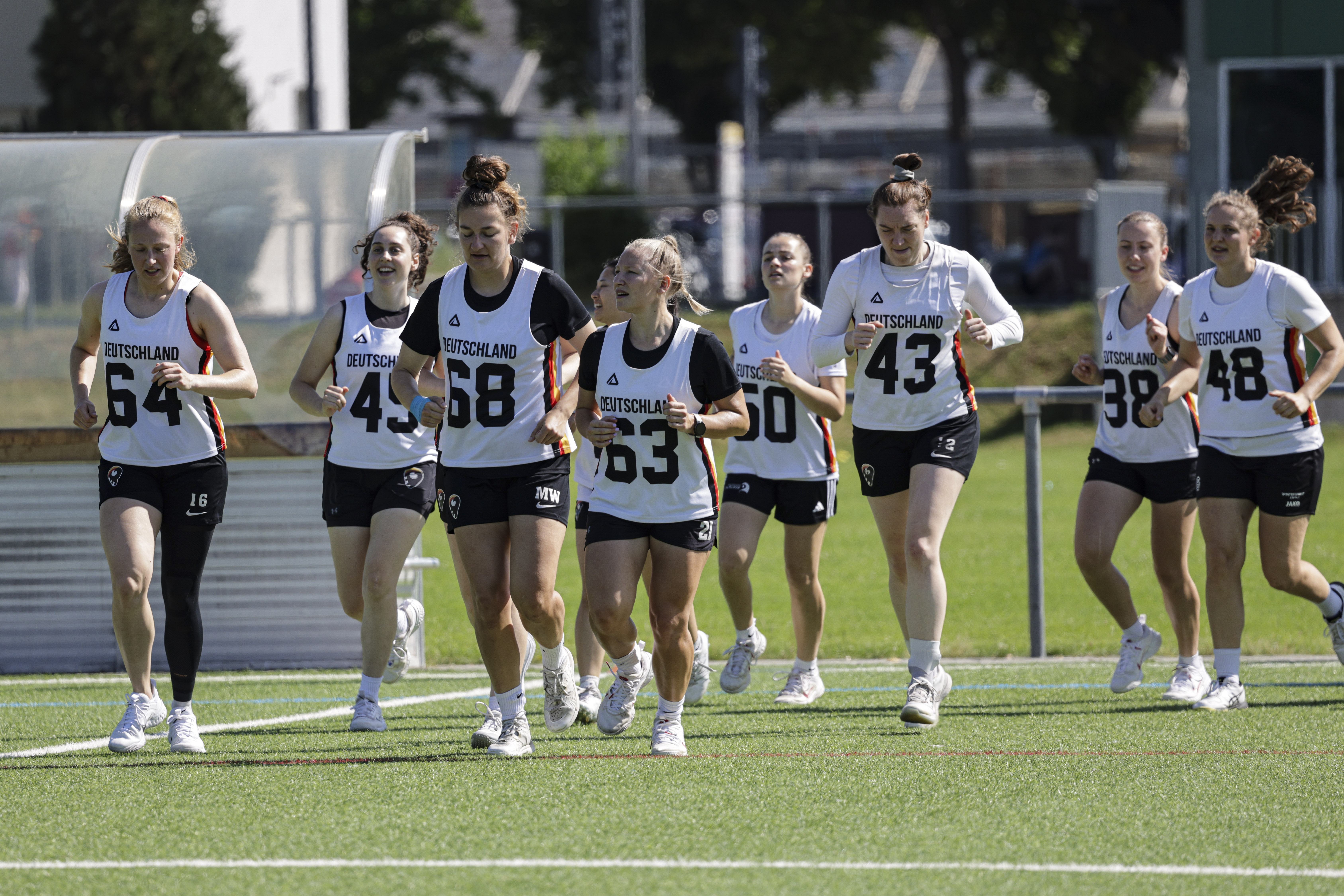 Spielerinnen der Lacrosse-Nationalmannschaft laufen in weißen Trikots und schwarzer Hose gemeinsam über das Spielfeld.