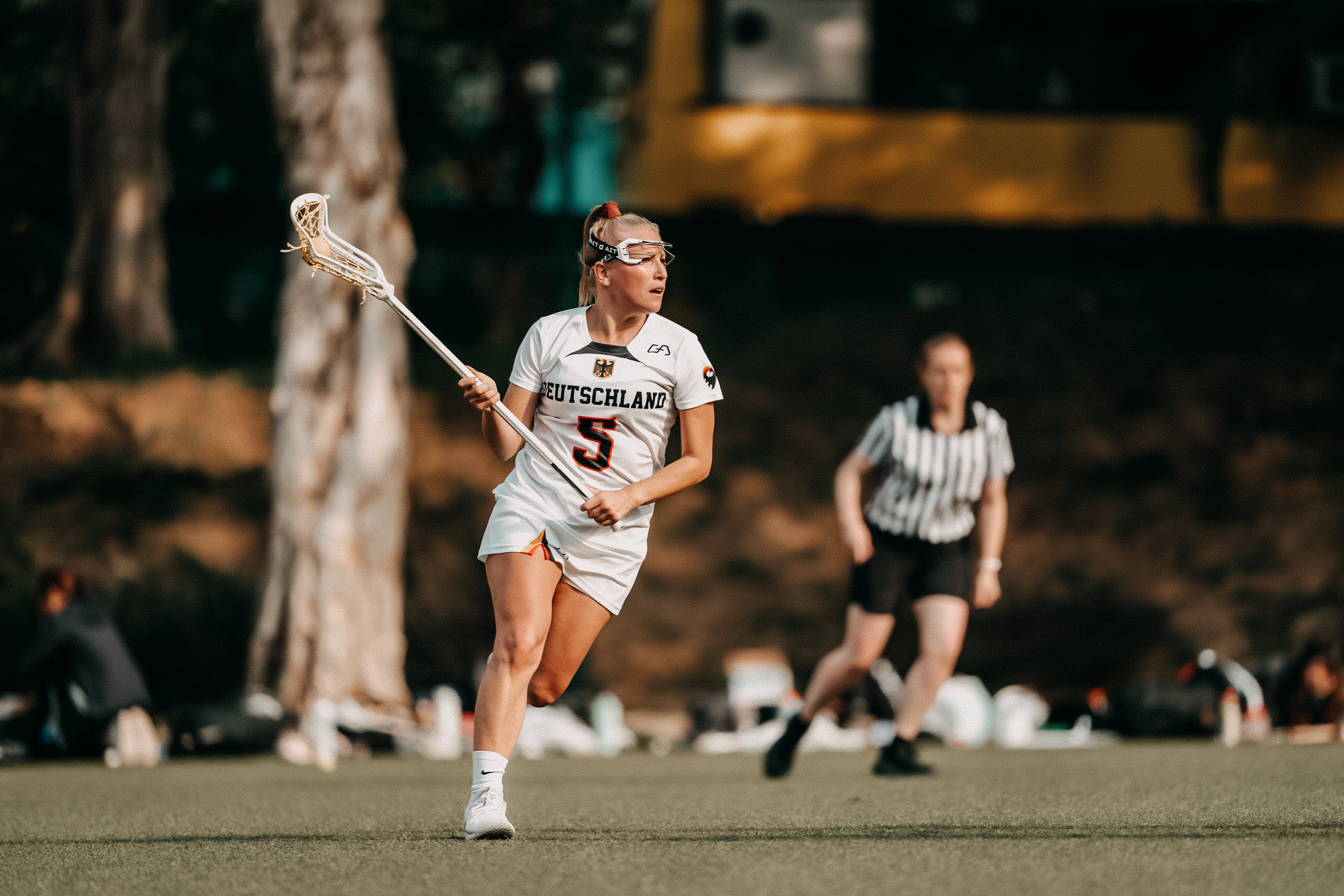 Lacrosse-Nationalspielerin Lena Johannsen läuft im weißen Deutschlandtrikot und mit Gesichtsschutz über das Spielfeld. Dabei hält sie ihren Lacrosse-Schläger in die Höhe.