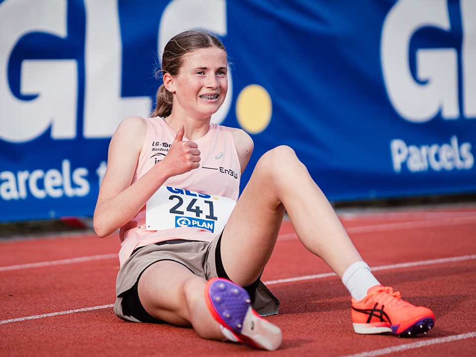 Julia Ehrle Auf dem Bild ist eine Person in Sportbekleidung auf einer Leichtathletikbahn zu sehen. Die Person sitzt auf dem Boden der Laufbahn und trägt einen Startnummernaufdruck, während im Hintergrund eine blaue Werbebande mit Sponsorenschriftzügen erkennbar ist. Die Aufnahme entstand im Umfeld eines Leichtathletikwettkampfs und rückt die Athletin beziehungsweise den Athleten in den Mittelpunkt.