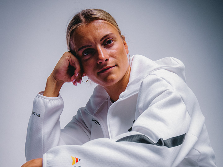 Auf dem Bild ist eine Person in einer weißen Sportjacke vor einem neutralen, hellen Hintergrund zu sehen. Die Person sitzt in einer entspannten Haltung und blickt in die Kamera, während die reduzierte Bildgestaltung den Fokus auf die sportliche Bekleidung und die Porträtaufnahme legt. Die Aufnahme vermittelt einen modernen und hochwertigen Eindruck.