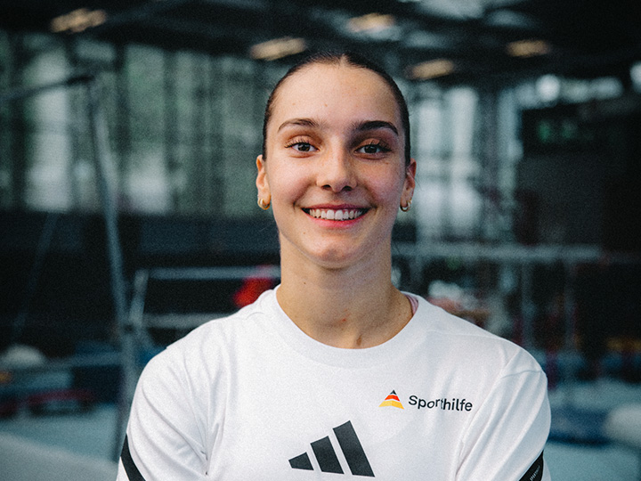 Die Aufnahme zeigt ein Porträt einer Person in Sportkleidung vor dem unscharfen Hintergrund einer Turnhalle. Die klare Bildkomposition und die helle Ausleuchtung lenken den Fokus auf die Person und unterstreichen den Kontext des Leistungssports.